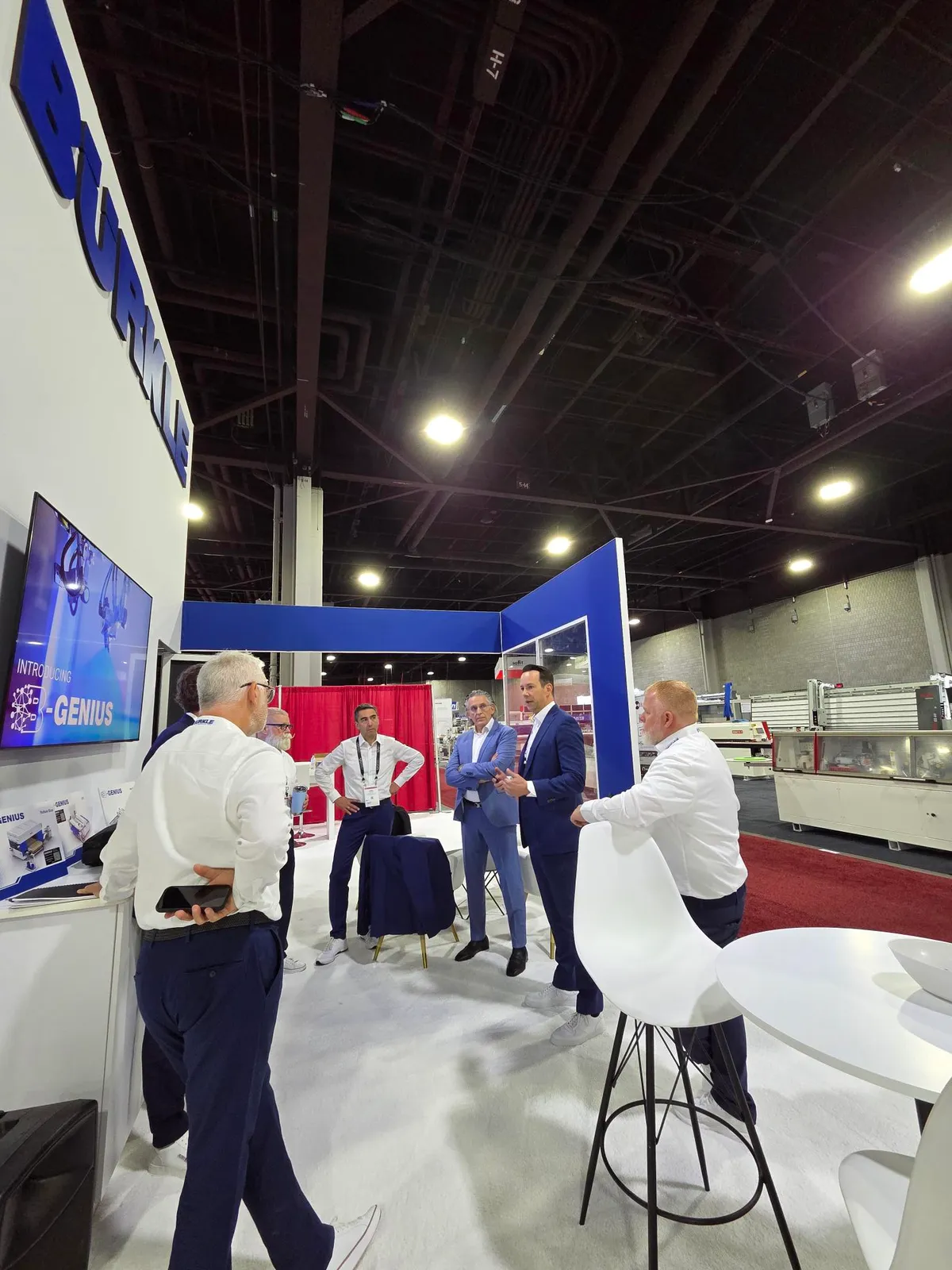 Visitors in conversation at the BÜRKLE booth at IWF Atlanta 2026, with a screen announcing "Introducing B-GENIUS" in the background